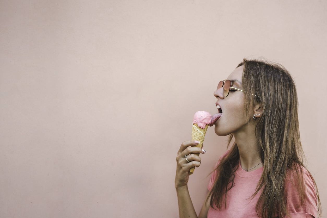 Beautiful, sexy model girl in sunglasses and pink t-shirt holding and licking tasty pink strawberry ice cream on pink background. Hot summer day.